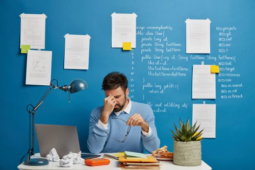Exhausted businessman rubs nose, takes off spectacles, suffers from eye strain and headache, has problems at work, sits in coworking space with laptop computer, blue wall with written notes. Exhausted businessman rubs nose, takes off spectacles, suffers from eye strain and headache, has problems at work, sits in coworking space with laptop computer, blue wall with written notes.