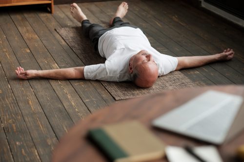 Senior caucasian man meditating on a wooden floor of living room and lying in Shavasana pose after practice. Yoga relax, meditating concept, close up